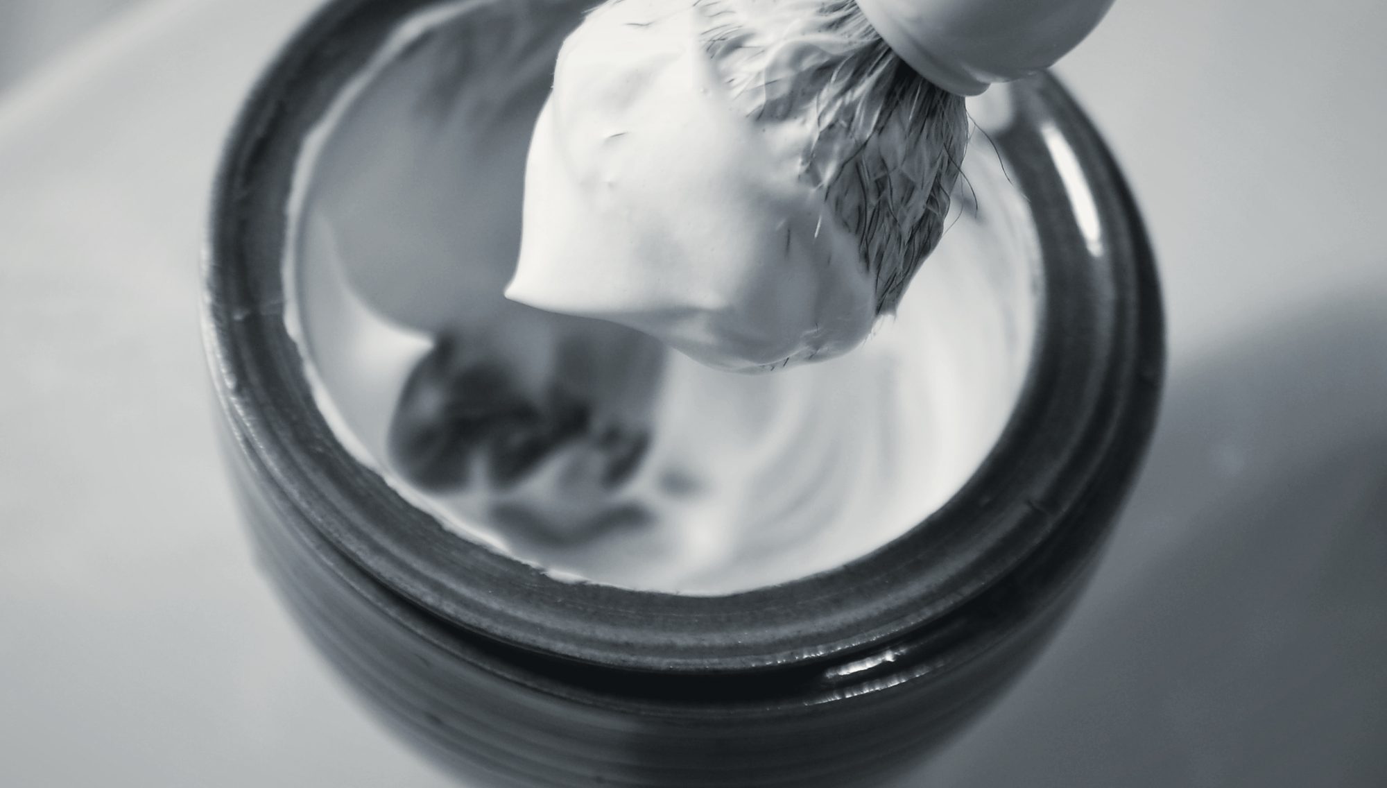 A shaving brush lifting thick lather out of a warmed stoneware bowl