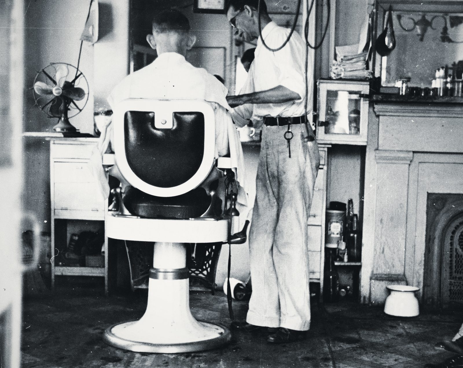 A 1935 barbershop: a barber in shirtsleeves works behind a white porcelain chair, a desk fan and bottles along the wall